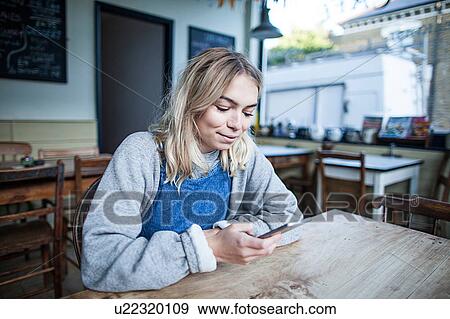 Young woman sitting in cafe, using smartphone, smiling View Large Photo Image Stock Photo - Young woman sitting in cafe, using smartphone, smiling. Fotosearch