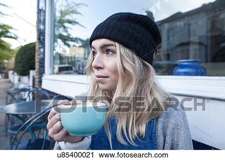 Stock Image - Young woman sitting outside cafe, drinking cup of tea. Fotosearch