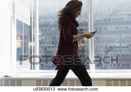 Stock Image - Young woman walking with mobile phone in front of window. Fotosearch