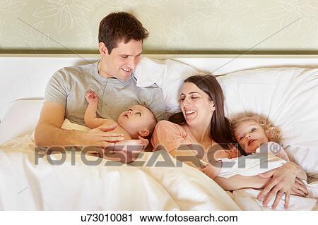 Stock Image - Baby girl and toddler sister huddled in bed with parents. Fotosearch