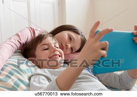 Stock Image - Boy and sister reclining on beanbag chair looking at digital tablet. Fotosearch
