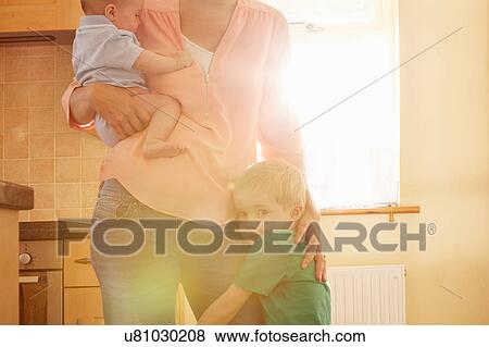 Stock Photo - Boy hugging mother's leg while she carries his baby brother in kitchen. Fotosearch