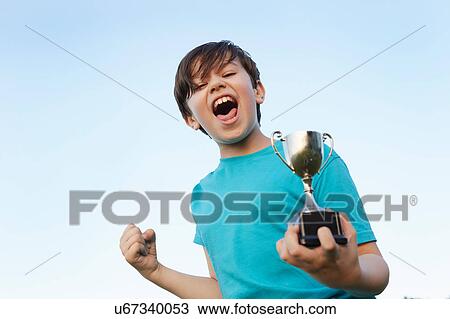 Stock Image - Boy making fist and holding sport trophy against blue sky. Fotosearch
