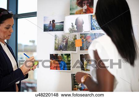 Stock Photo - Colleagues in office discussing photographs on wall. Fotosearch