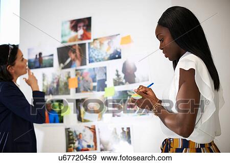 Stock Photo - Colleagues in office discussing photographs on wall. Fotosearch