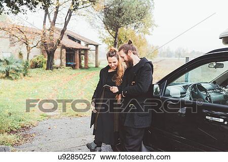 Stock Photo - Couple leaning against car looking at smartphone. Fotosearch