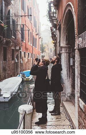 Couple photographing building exteriors from canal waterfront, Venice, Italy View Large Photo Image Stock Photograph - Couple photographing building exteriors from canal waterfront, Venice, Italy. Fotosearch