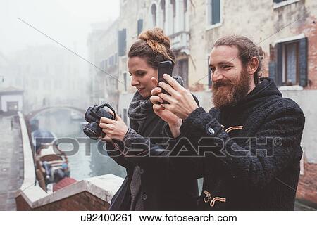 Stock Image - Couple photographing on misty canal waterfront, Venice, Italy. Fotosearch
