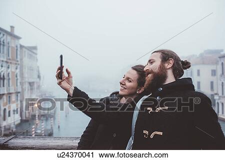 Stock Image - Couple taking smartphone selfie over misty canal, Venice, Italy. Fotosearch