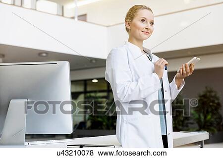 Doctor standing beside computer, holding smartphone View Large Photo Image Stock Photo - Doctor standing beside computer, holding smartphone. Fotosearch