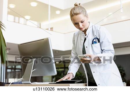 Doctor standing beside computer, holding open diary, looking at smartphone View Large Photo Image Stock Photograph - Doctor standing beside computer, holding open diary, looking at smartphone. Fotosearch