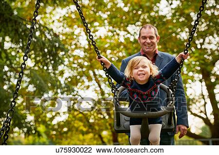 Stock Image - Father pushing daughter on playground swing. Fotosearch