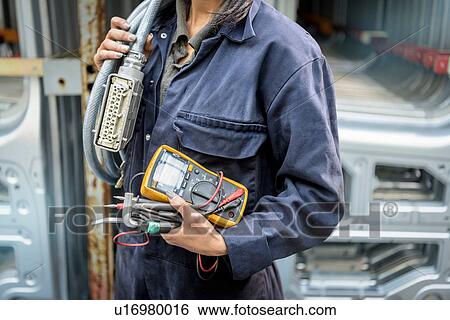 Stock Photograph - Female apprentice electrician holding equipment in car factory, close up. Fotosearch