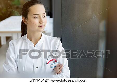 Stock Photo - Female doctor looking out of window, arm folded. Fotosearch