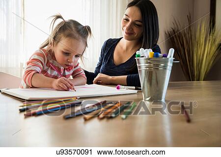 Stock Image - Female toddler at table with mother drawing in sketchbook. Fotosearch