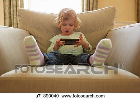 Stock Image - Female toddler sitting on armchair playing hand held computer game. Fotosearch