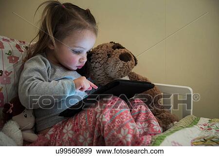 Stock Photo - Female toddler sitting up in bed using touchscreen on digital tablet. Fotosearch