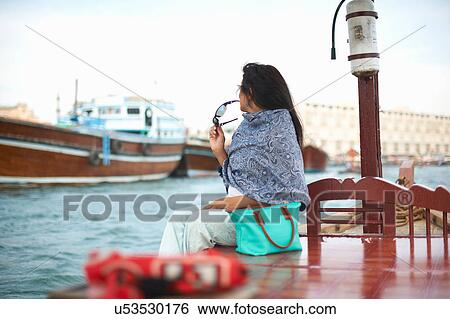 Stock Photograph - Female tourist sitting on waterfront watching boats on creek, Dubai, United Arab Emirates. Fotosearch