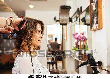 Picture - Hairdresser using curling tongs on customer's long brown hair in salon. Fotosearch