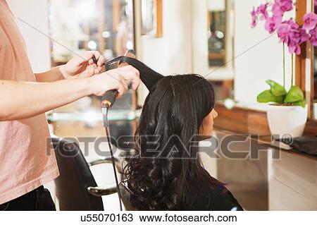 Stock Image - Hairdresser using curling tongs on customers long black hair in salon. Fotosearch