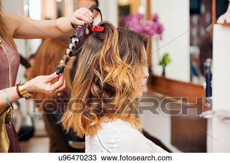 Stock Image - Hairdresser using curling tongs on customer's long brown hair in salon. Fotosearch