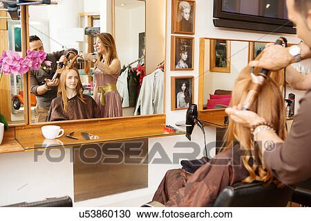 Stock Image - Hairdressers using curling tongs on customers long red hair in salon. Fotosearch