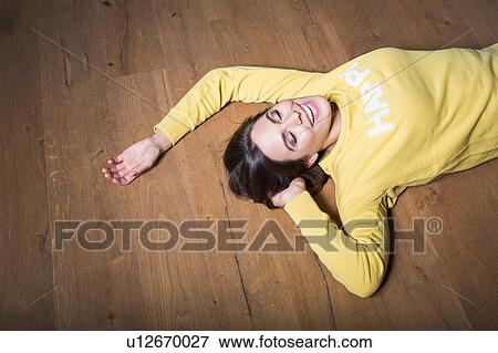 Stock Photo - Happy young woman in yellow sweatshirt lying on wooden floor. Fotosearch