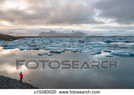 Stock Image - High angle view of tourist looking out at icebergs on glacial lagoon, Jokulsarlon, Iceland. Fotosearch