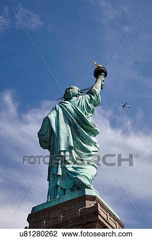 Stock Image - Low angle view of Statue of Liberty and airplane in blue sky, New York city, USA. Fotosearch