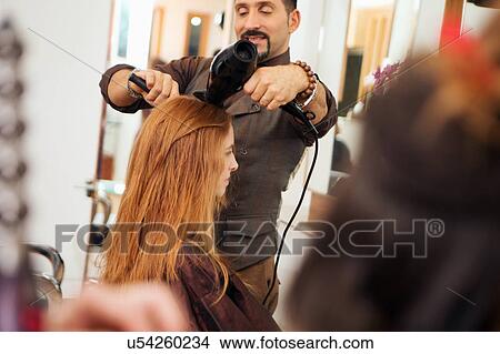 Picture - Male hairdresser blow drying customer's red hair in hair salon. Fotosearch