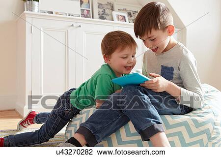 Stock Image - Male toddler and brother on beanbag chair looking at digital tablet. Fotosearch