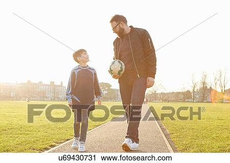Stock Image - Man and football player son strolling and talking in park. Fotosearch