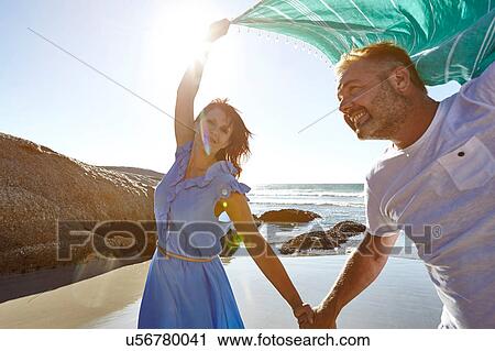 Stock Image - Mature couple holding hands on beach, woman holding sheer scarf in air. Fotosearch