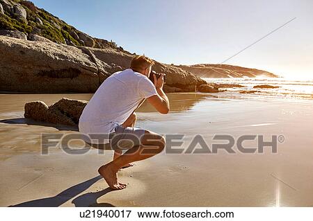 Stock Photo - Mature man crouching on beach, photographing view, Cape Town, South Africa. Fotosearch