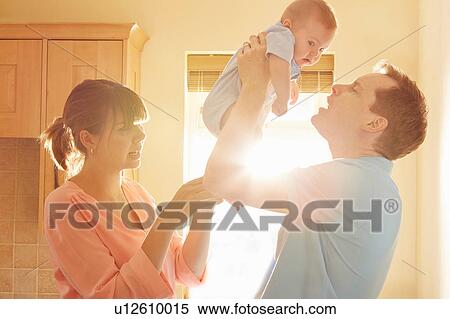 Stock Photography - Mature man with wife holding up baby son in sunlit kitchen. Fotosearch