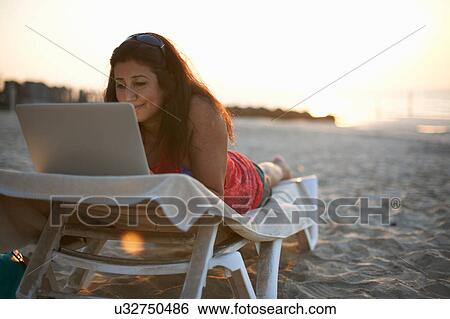 Stock Photograph - Mature woman lying on beach sun lounger looking at laptop, Dubai, United Arab Emirates. Fotosearch