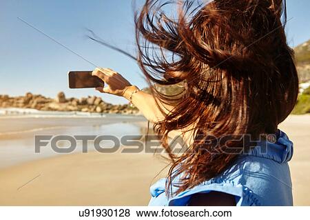 Stock Photo - Mature woman on beach, taking selfie, using smartphone, rear view. Fotosearch