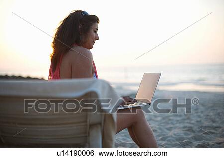 Stock Photograph - Mature woman sitting on beach sun lounger looking at laptop, Dubai, United Arab Emirates. Fotosearch