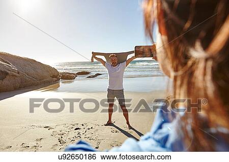 Stock Photograph - Mature woman taking photograph of man on beach, Cape Town, South Africa. Fotosearch