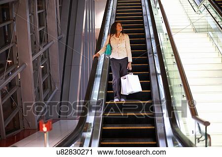 Stock Image - Mature woman with shopping bags moving down escalator in shopping mall, Dubai, United Arab Emirates. Fotosearch