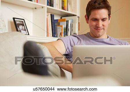 Picture - Mid adult man sitting on sofa looking at laptop. Fotosearch