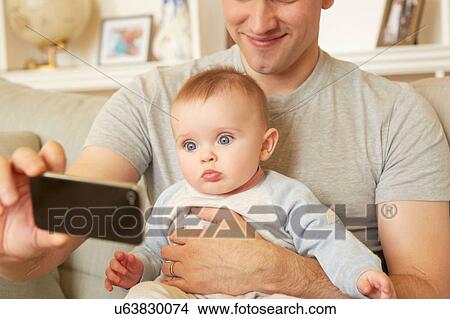 Picture - Mid adult man taking smartphone selfie with baby daughter on sofa. Fotosearch