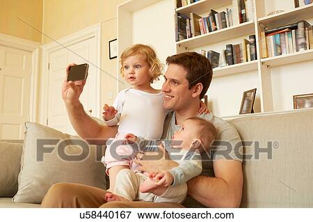 Stock Photo - Mid adult man taking smartphone selfie with toddler and baby daughter on sofa. Fotosearch