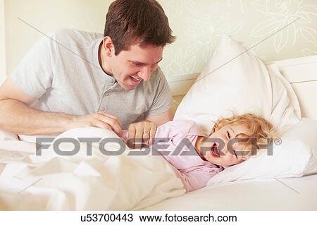 Stock Image - Mid adult man tickling toddler daughter in bed. Fotosearch