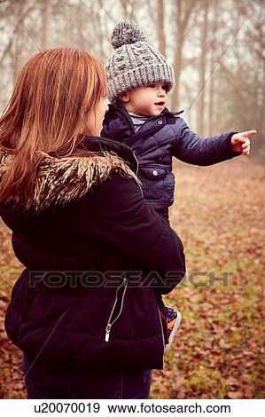 Stock Photo - Mid adult woman carrying toddler son pointing in autumn forest. Fotosearch
