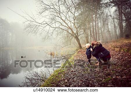 Picture - Mid adult woman crouching with toddler son on misty riverbank. Fotosearch