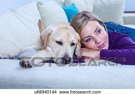 Picture - Mid adult woman lying on sofa with her labrador dog. Fotosearch