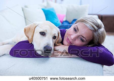 Stock Image - Mid adult woman lying on sofa with her labrador dog. Fotosearch