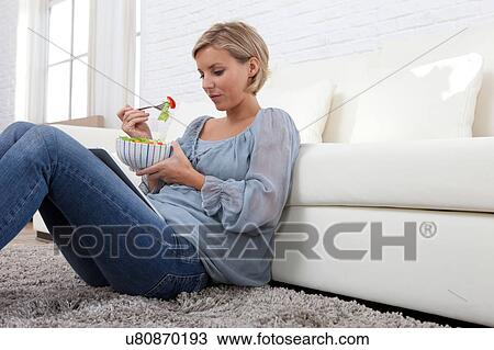 Stock Image - Mid adult woman sitting on rug looking at digital tablet and eating salad. Fotosearch