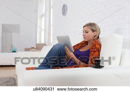 Stock Image - Mid adult woman sitting on sofa looking at digital tablet. Fotosearch